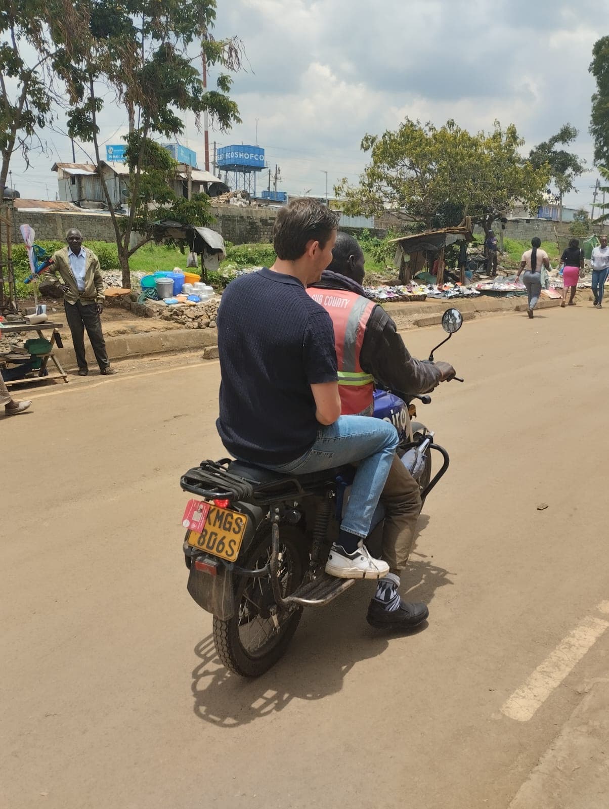 A guest riding pillion on a boda boda down a Kibera street, the rider in a high visibility vest