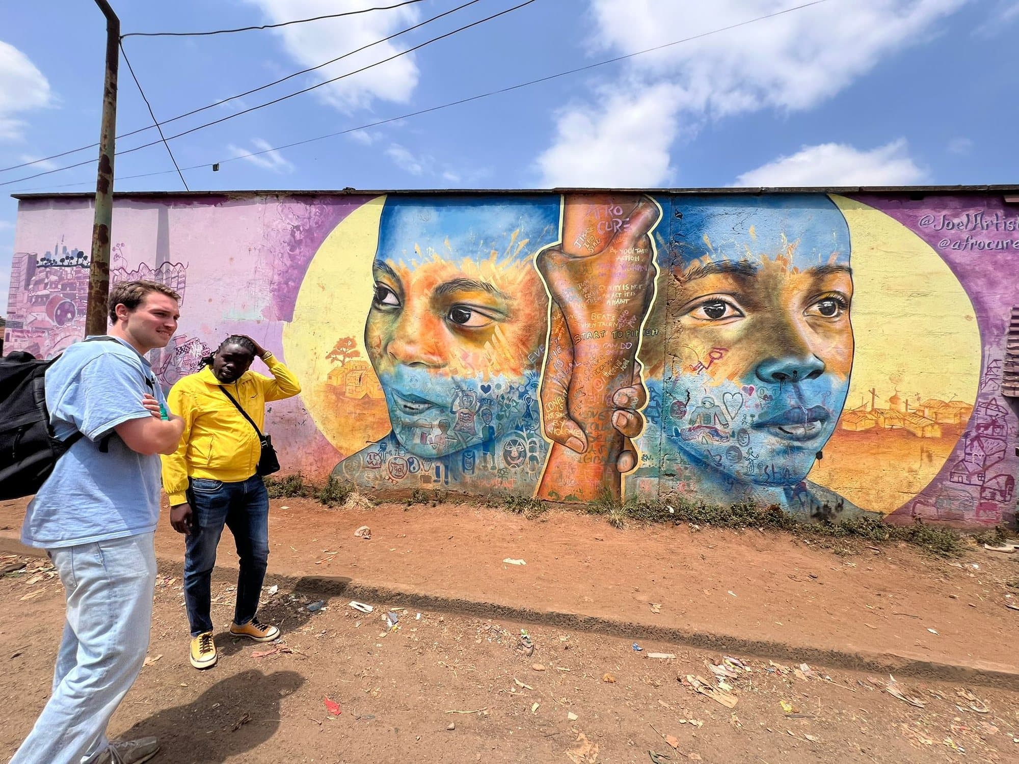 A painted wall in Kibera showing two large faces holding hands, with Sammy and a guest walking past it